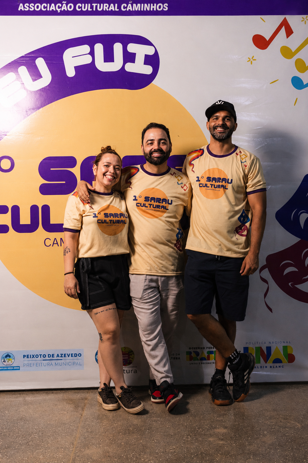 Três integrantes da equipe organizadora do 1º Sarau Cultural sorrindo em frente ao banner oficial
